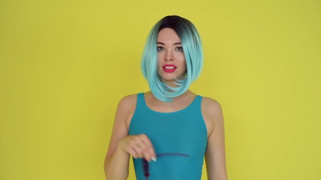 Beautiful girl in a blue wig and bikini posing on a yellow background. Woman with artificial hair and a swimsuit. heart-shaped sunglasses