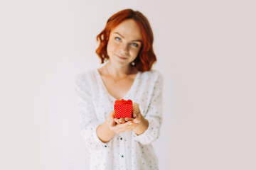 Young birthday girl holding a small red gift box, isolated on white background. Attractive female model in white clothes. Focus on a box.