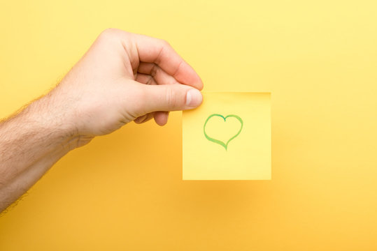 cropped view of man holding sticky note with heart on yellow background