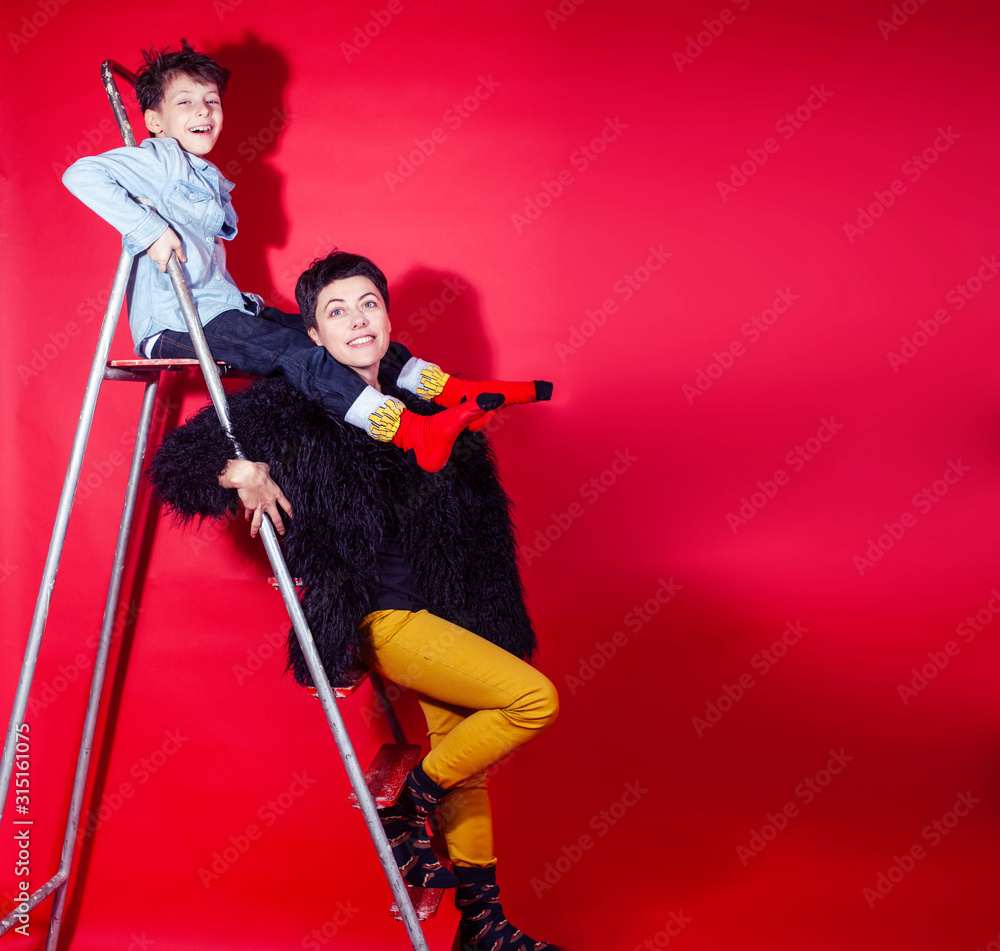 Wall mural pretty young mother with little son on ladder on red background posing cheerful happy smiling, lifes