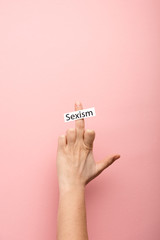 cropped view of woman showing middle finger and card with sexism lettering on pink background