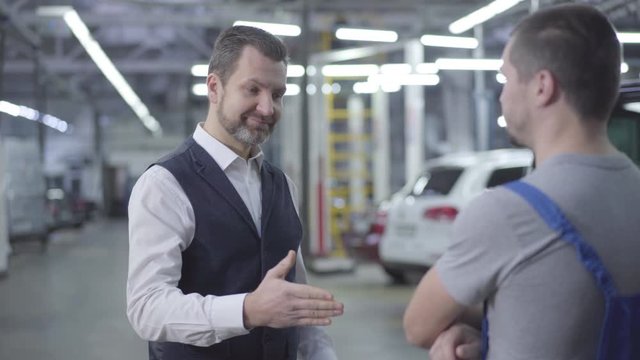 Portrait of adult bearded Caucasian man in elegant outfit taking car keys from auto mechanic and shaking his hand. Owner taking back fixed car from repair shop. Warranty service, car repair.