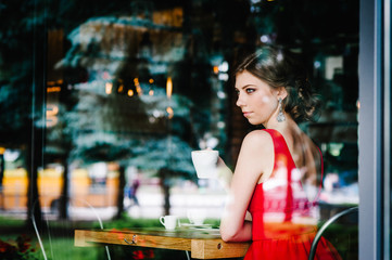 Fototapeta na wymiar Beautiful serious European brunette girl in an evening red dress sitting at the window with a cup of coffee at the cafe. Looking at the side through the window.