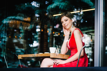 Beautiful happy European brunette girl in an evening red dress sitting at the window with a cup of coffee at the cafe. Looking in the camera through the window.