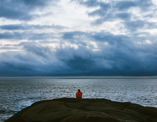 A lonely man in a yellow T-shirt sits on the edge of a huge stone seeing off the sunset. Dreamy and romantic concept. Figure of a man against the backdrop of the ocean and dramatic clouds gathering.
