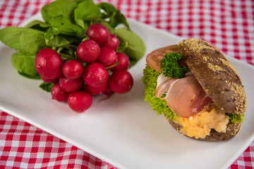 on a white plate lies a sliced bread roll filled with sausage, cheese and egg and as decoration a bunch of radishes