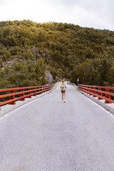 Back view of young blonde woman with backpack walking on the road over a bridge near the mountain. Travel and adventure concept. Traveler in the middle of woodland. Travel alone