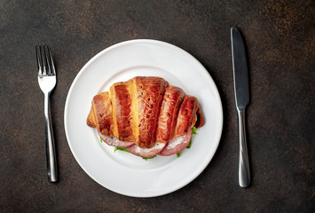 croissant - a sandwich with sausage and cheese in a white plate with a fork and a knife on a stone background with copy space for your text