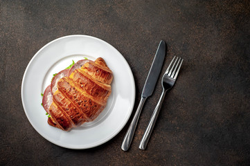croissant - a sandwich with sausage and cheese in a white plate with a fork and a knife on a stone background with copy space for your text