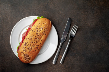 large panini sandwich with sausage and cheese in a white plate with a knife and fork on a stone background with copy space for your text