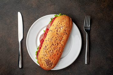 large panini sandwich with sausage and cheese in a white plate with a knife and fork on a stone background with copy space for your text