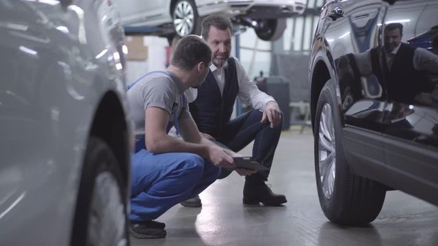 Two Caucasian adult men sitting on hunkers next to car and talking. Professional repair man in blue robe consulting client regarding repair of his car's wheels. Repair services, car business.