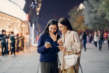 two girlfriends using their cellphone while exploring a new city at night. Two friends searching for information on their cellphone as they are exloring Beijing at night. Girlfriends pointing at an