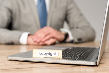 selective focus of wooden block with word copyright on laptop near businessman sitting at desk isolated on grey