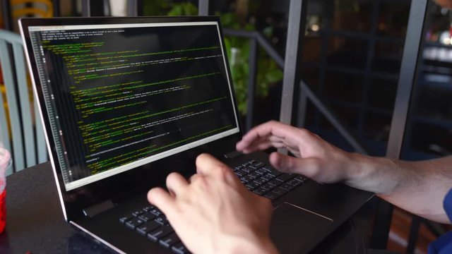 Back view over the shoulder shot of developer programmer with laptop. Program code and script data on the screen. Young freelancer in glasses working on project in cafe.