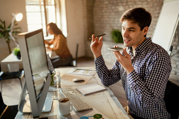 Young businessman recording voice message on cell phone while working on a computer.