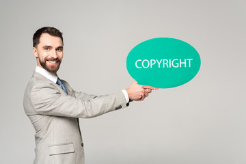 handsome businessman holding speech bubble with word copyright and smiling at camera isolated on grey