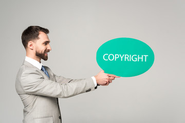 side view of smiling businessman holding thought bubble with copyright inscription isolated on grey