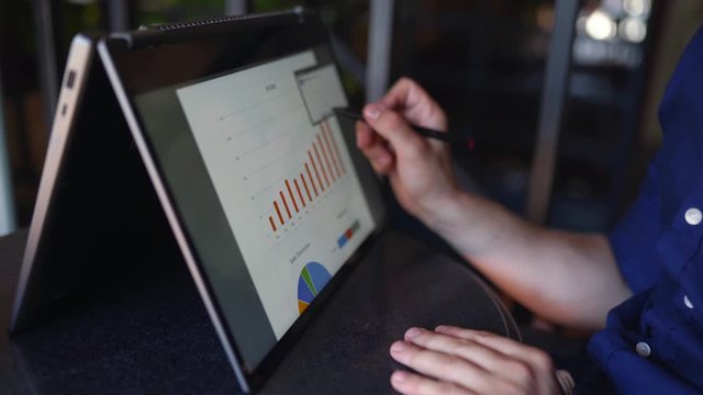 Businessman makes a presentation using stylus and convertible laptop with touchscreen in tent mode. Freelancer works with 2 in 1 transformer notebook with touch display. Pen pointing at graphic chart.