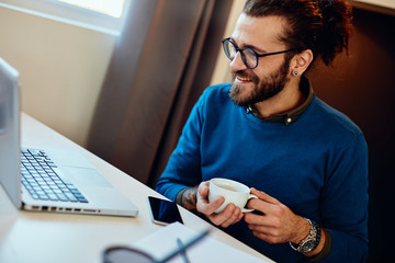 Charming caucasian bearded hipster sitting in his office, looking at laptop, holding coffee and taking a break.