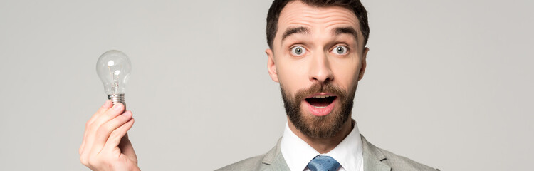 Obraz premium panoramic shot of excited businessman holding light bulb and looking at camera isolated on grey