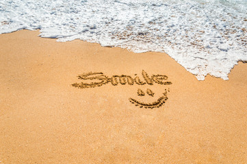 Hand written text smile and symbol on the golden beach sand with coming wave