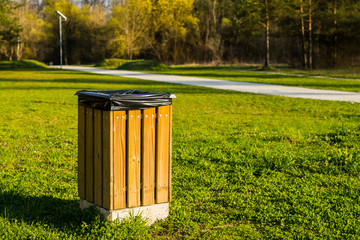 A modern wooden trash can or bin in a park with a black plastic bag visible inside on a sunny day. Walkway or path for pedestrians in the background.