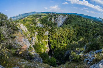 Unesco world heritage site Skocjanske jame taken with a wide fish eye view. Looking towards the deep gorge with cave entrance and walking paths. Village of Skocjan in the background. © Anze