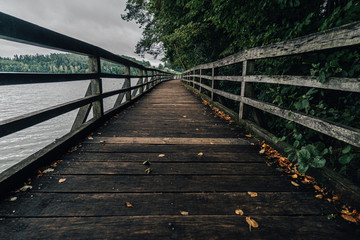 Fototapeta premium Wooden walkway or bridge on the side of a lake. Old wooden walkway on the banks of a lake. Autumn view of a wooden path and a lake.