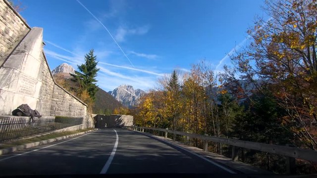 Inside car dashboard point of view forward moving driving on Slovenia road in valley between Alps and colorful trees, passing Fort Predil ruins with view of Mangart mountain in background