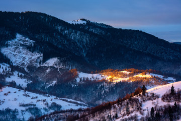 winter countryside scenery at dawn. landscape with spot of first light on snow covered hill. dark coniferous distant forest in shade of the mountain. cold weather.