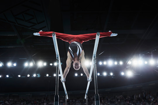 Male gymnast performing upside-down splits on parallel bars in arena