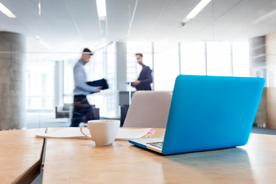 Laptop coffee cup on conference room table with businessmen walking 