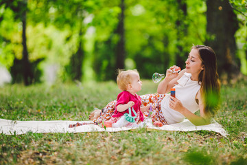 Smiling woman play and sit on green grass in park, rest and hug hold soap bubble blower with little...