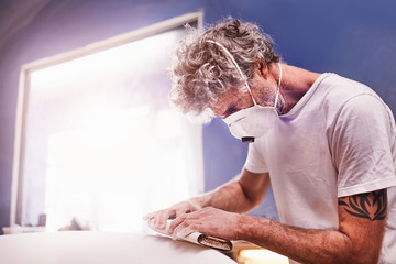 Focused man wearing protective mask sanding surfboard in workshop