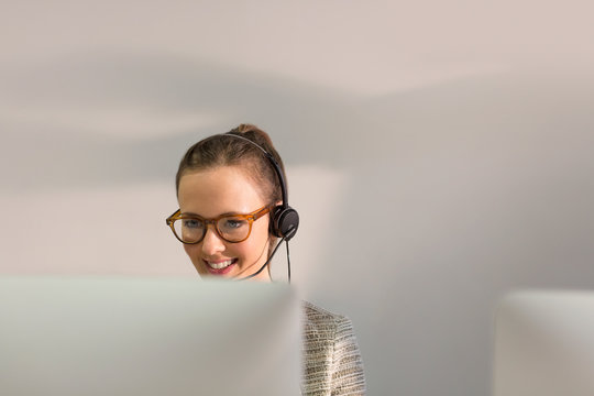 Smiling female telemarketer wearing headset talking on telephone at computer in office