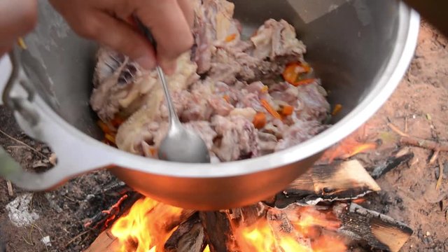 Man hand mix roasted vegetables and chicken parts with spoon in big metal cauldron, hanging over the fire. Outdoor food in camp. Touristic lagman. Process of cooking
