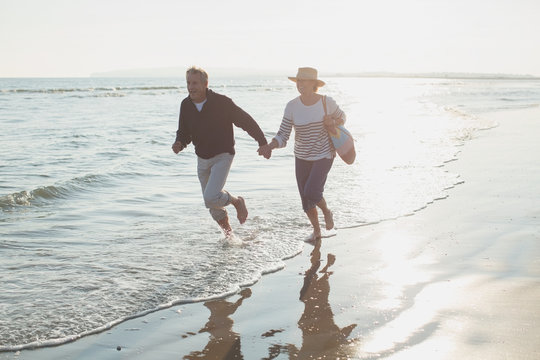 Playful mature couple holding hands and running in sunny ocean surf