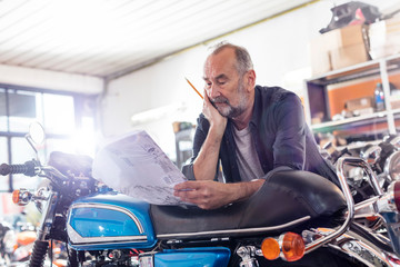 Focused senior male motorcycle mechanic reviewing manual in workshop