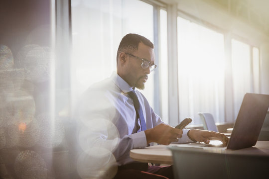 Serious Businessman Texting With Cell Phone At Laptop In Office