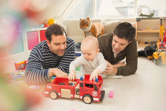 Male gay parents and baby son playing with fire engine toy in playroom