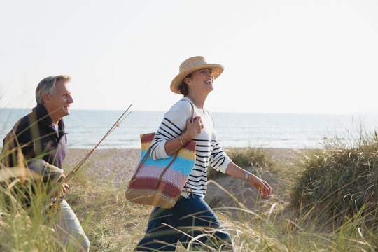 Mature couple with fishing rod walking in sunny beach grass