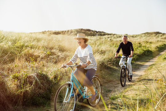 Playful mature couple riding bicycles on sunny beach grass path