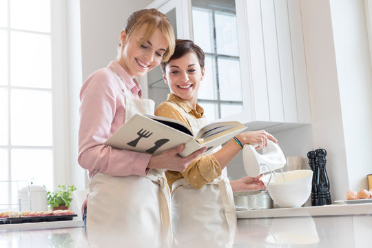 Female caterers with cookbook baking in kitchen, using electric hmixer