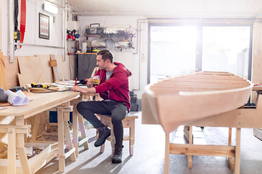 Male carpenter drinking tea working at laptop on workbench in workshop