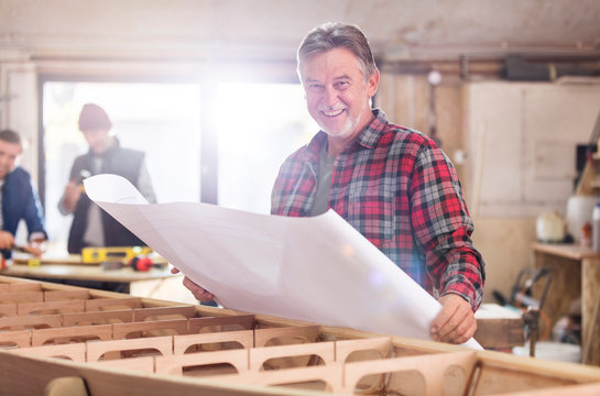 Portrait smiling male carpenter reviewing plans at wood boat in workshop