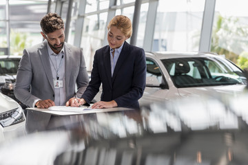 Car salesman watching female customer signing financial contract paperwork in car dealership showroom