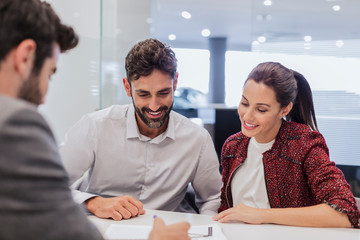 Car salesman explaining financial contract paperwork to couple customers in car dealership office