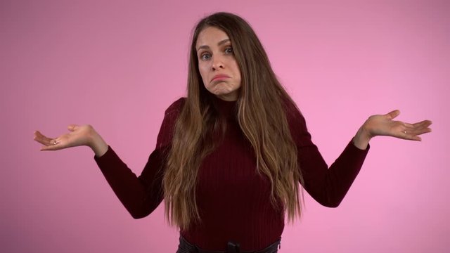 Attractive brunette woman throwing hands wide and shaking shoulders in despair. Displeased woman showing perplexity emotions feeling misunderstanding. Confused woman showing helpless gesture over pink