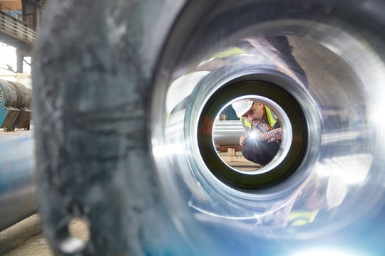 Male engineer with flashlight examining steel pipe in factory